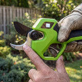 A close-up of a person's hands holding a green and black pruning shears tool outdoors in a garden setting, with one hand wearing a work glove.