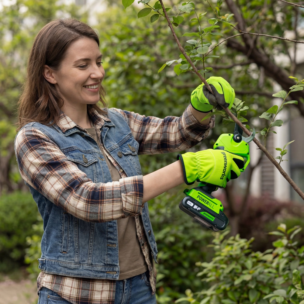 A woman wearing a denim vest and plaid shirt uses a cordless hedge trimmer with bright green gloves to prune a leafy green bush in a garden.