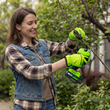 A woman wearing a denim vest and plaid shirt uses a cordless hedge trimmer with bright green gloves to prune a leafy green bush in a garden.