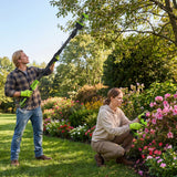 A man and a woman in a lush garden use cordless garden tools. The man stands on the grass holding a long-reach hedge trimmer, while the woman crouches beside pink flowers using a handheld edger.